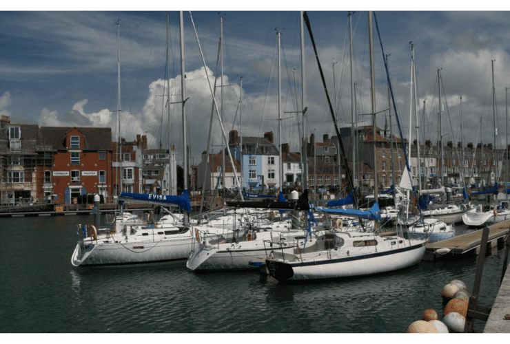 Several sailboats docked in a marina with a row of townhouses and cloudy sky in the background.