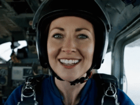 A woman wearing a helmet and parachute gear smiles inside an aircraft cockpit.