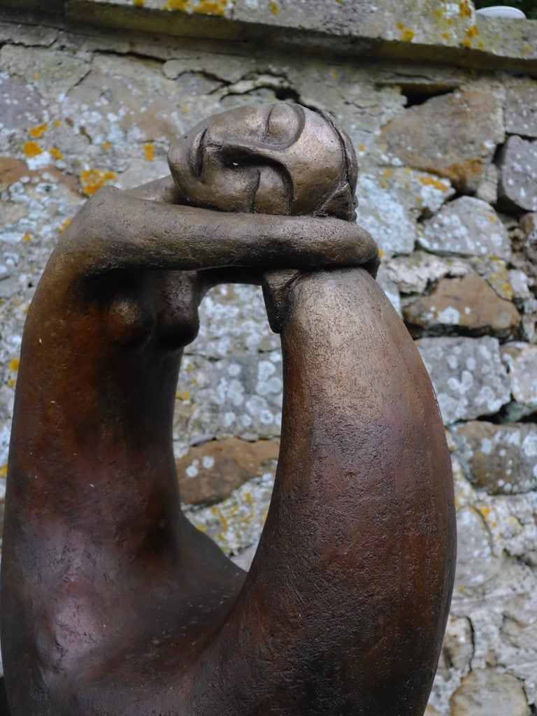 Bronze abstract sculpture of a person resting their head on crossed arms in front of a stone wall.