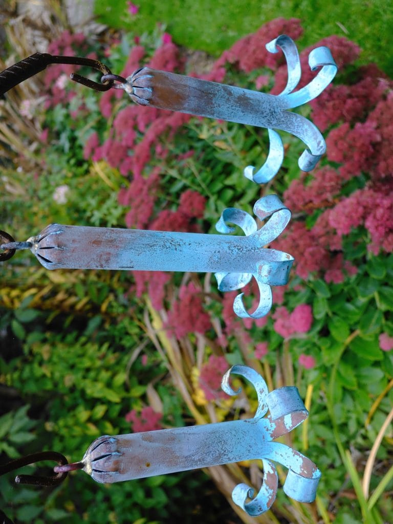 Three blue, weathered metal hooks are hanging outdoors with pink flowers and green foliage in the background.
