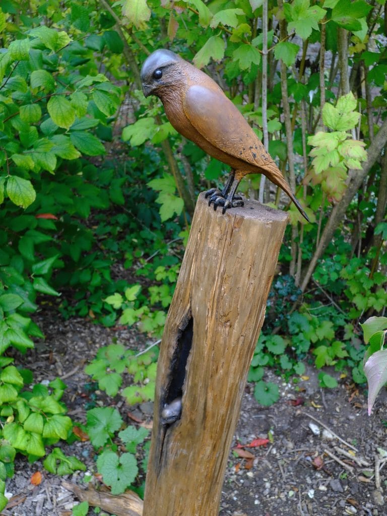 Bronze bird sculpture perched on a wooden post surrounded by green foliage.