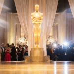 Large Oscar statue on stage, surrounded by elegantly dressed people and draped curtains at an awards event.