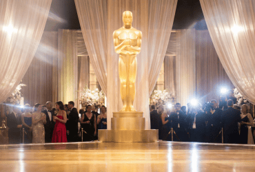 Large Oscar statue on stage, surrounded by elegantly dressed people and draped curtains at an awards event.