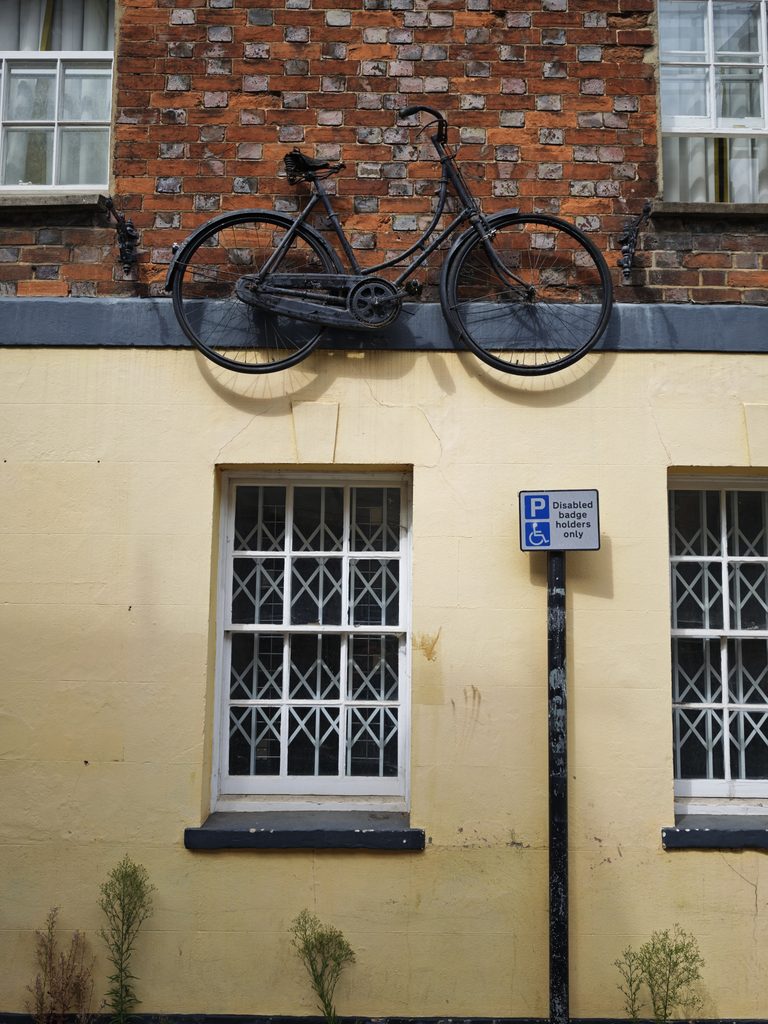 A black bicycle is mounted on a wall above a window, next to a disabled parking sign on a yellow building.