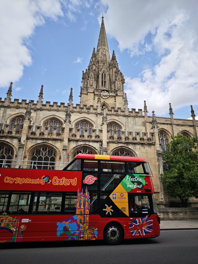 Red double-decker sightseeing bus passes in front of a historic stone church with a tall spire in Oxford.