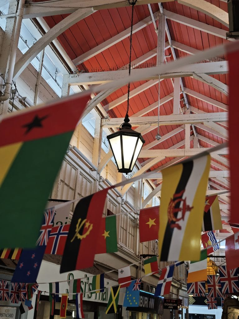 Colorful international flags hang from the ceiling of a market with a black lantern and red roof beams above.