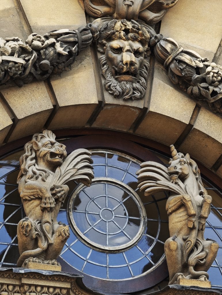 Stone lion and unicorn statues flank a round window below a sculpted lion head on an ornate building facade.