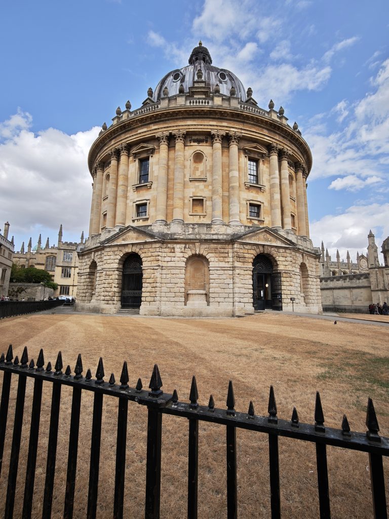 A large, round stone building with columns and a dome, seen behind a black metal fence on a cloudy day.