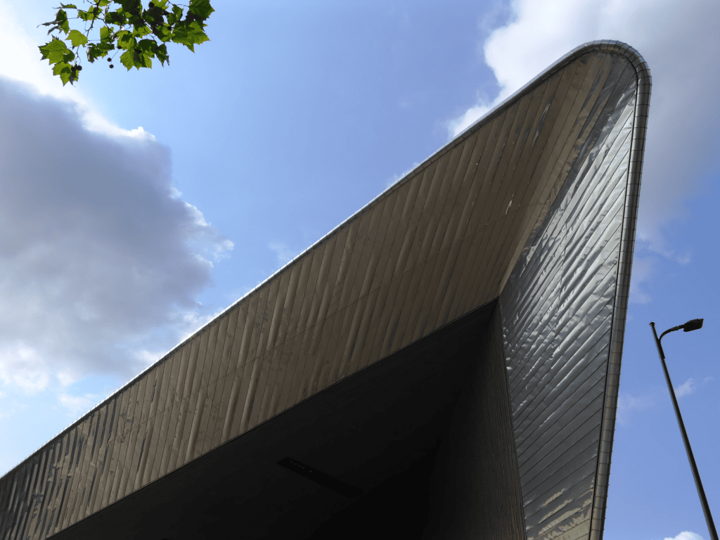 Modern building with a pointed metallic roof against a blue sky with clouds and a streetlight.