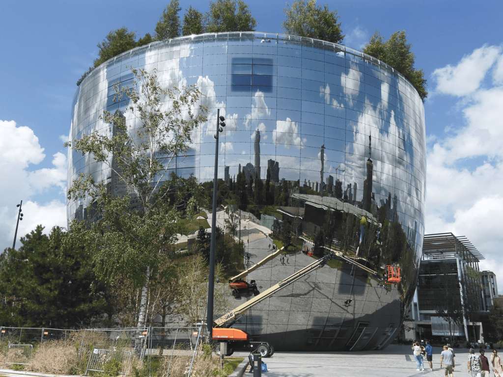 Circular glass building with mirrored surface reflecting trees, skyline, and construction equipment under partly cloudy sky.