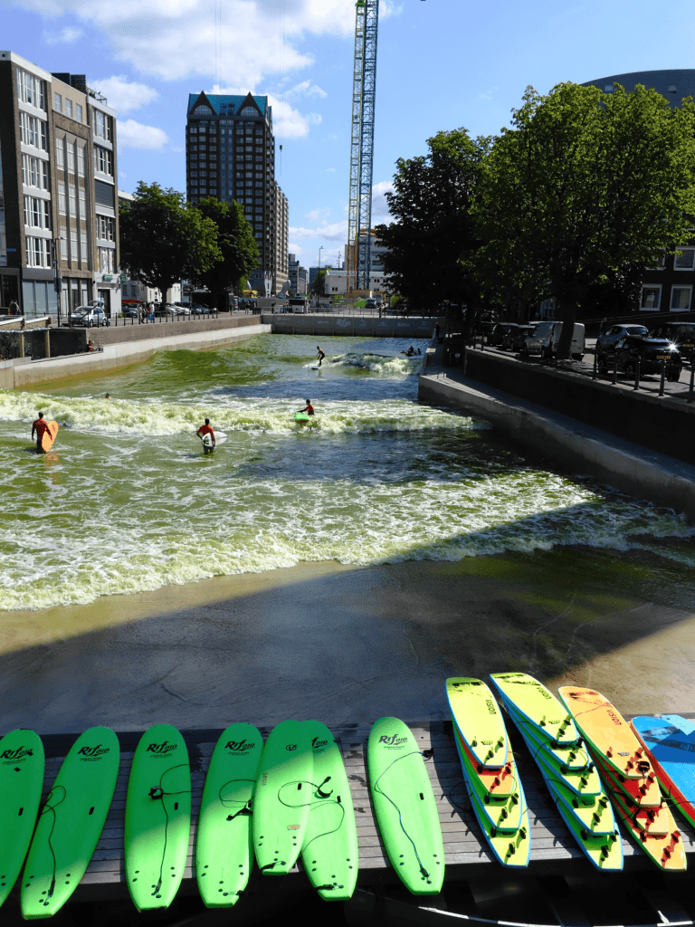 Several bright surfboards are lined up beside an urban artificial wave pool with people surfing in the background.