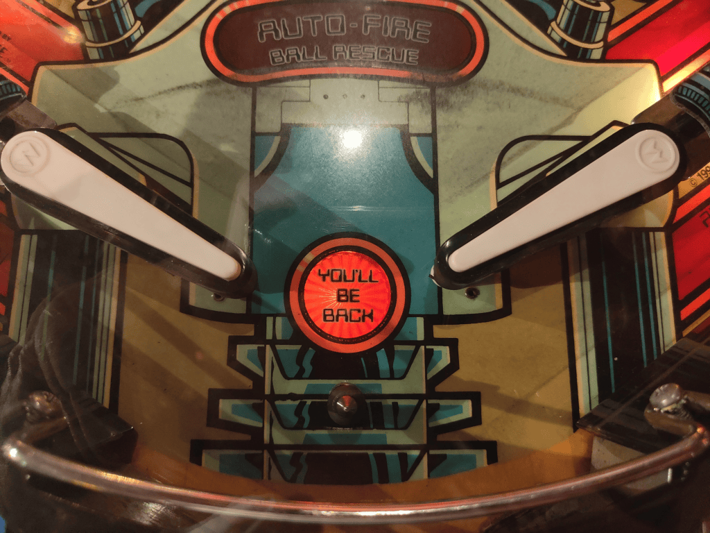 Close-up of a pinball machine playfield with the phrase “YOU’LL BE BACK” in a red circle between two flippers.