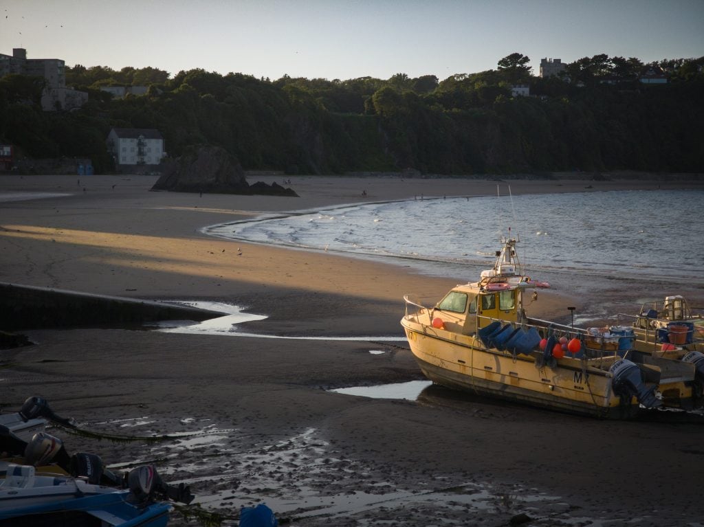 A yellow boat is beached on sand near the shoreline, with other boats and buildings visible in the background.