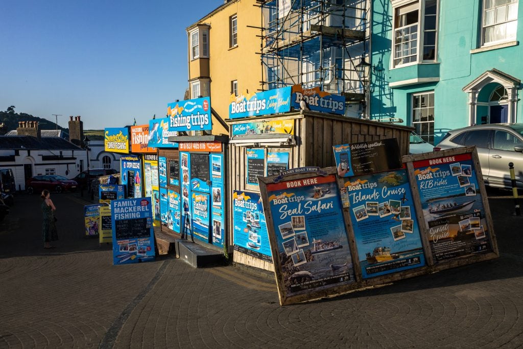 Outdoor kiosk with colorful signs advertising boat trips, fishing tours, and island camps in a sunny urban area.
