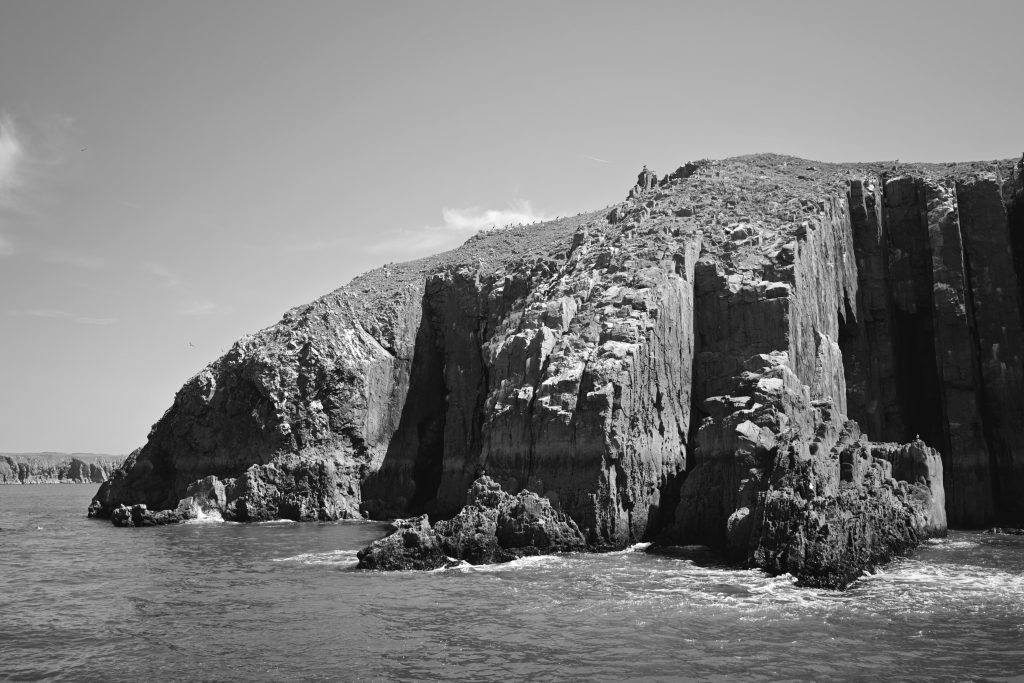 Rocky seaside cliffs rise from the water under a clear sky, with waves breaking at their base.