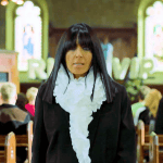 A woman in black stands in a church with people seated behind her and stained glass windows in the background.