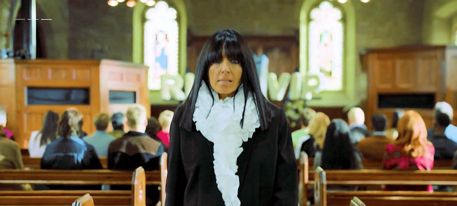 A woman in black stands in a church with people seated behind her and stained glass windows in the background.