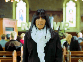 A woman in black stands in a church with people seated behind her and stained glass windows in the background.